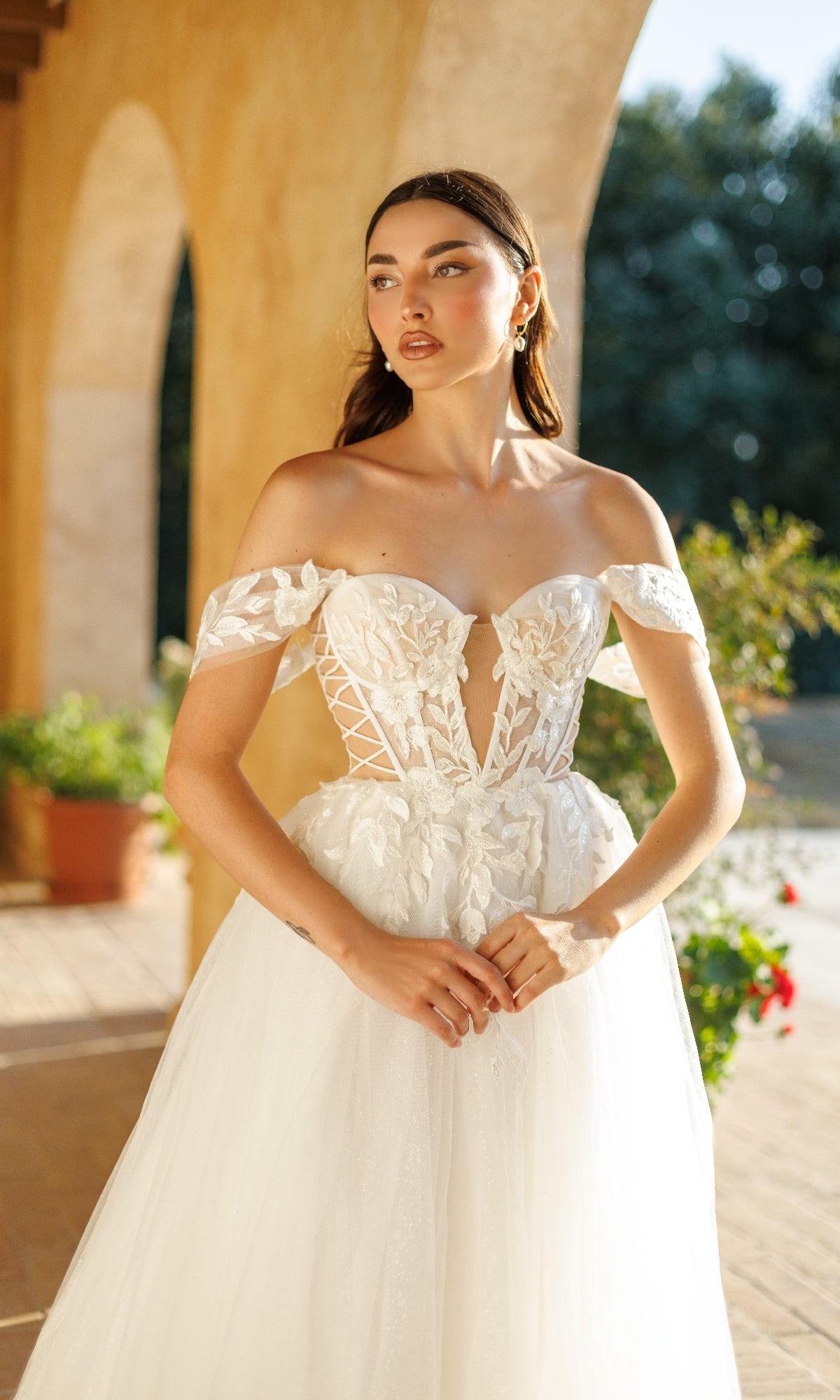 A woman stands outdoors in natural light, wearing the Long Formal Dress: Tina Holly TW480, an off-shoulder white bridal gown with a corset bodice and lace floral appliqué details.